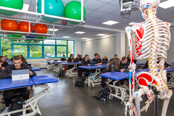 Schüler sitzen im Klassenraum an Tablets. Vorne im Bild steht ein Skelett